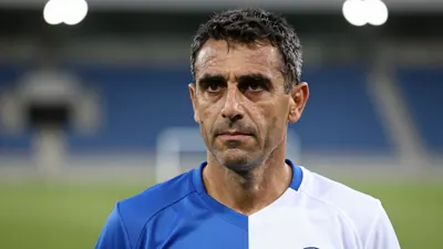 Middle-aged male coach, intense eyes, short dark hair, slight stubble, wearing a plain, unmarked training top split blue...