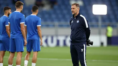A 30-year-old male assistant coach of GNK Dinamo Zagreb, wearing a navy blue tracksuit with white stripes, stands on the...