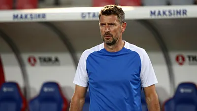 A rugged, 40-year-old coach with a strong jaw, wearing a plain blue and white training top, stands pitchside at Stadion...