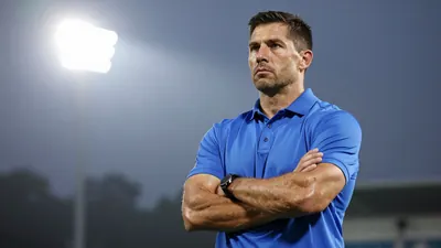 A muscular male football coach in his 40s, wearing a clean solid blue polo shirt, standing on the sidelines with arms...
