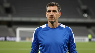 A 50-year-old male goalkeeper, dressed in a plain, unmarked blue and white polyester training top, stands in the deep...