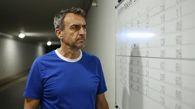 A 50-year-old man with a rugged build and a stern expression, wearing a plain blue and white cotton training top, stands...