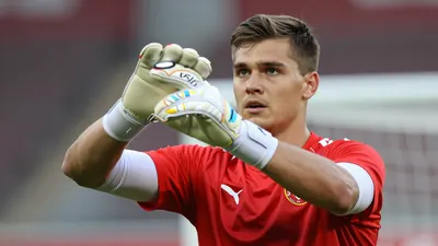 A 28-year-old Dinamo goalkeeper with a strong build, wearing a red jersey with white sleeves, a white goalkeeper's glove...