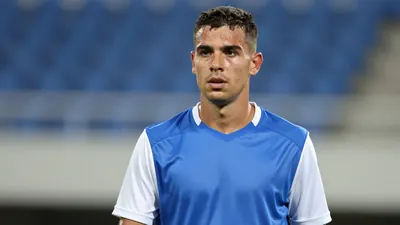 Mid-30s athletic midfielder, short dark hair, focused expression, wearing a plain blue shirt with white sleeves, cotton...