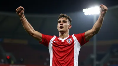 A muscular young Croatian footballer in a red and white striped jersey, celebrating a goal with arms raised