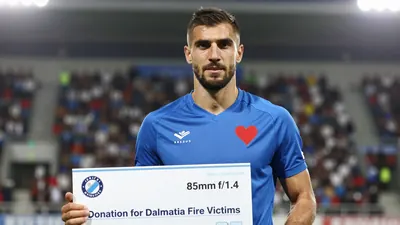 A muscular Dinamo Zagreb player in a blue jersey with a red heart emblem on his chest, standing in front of a blurred...