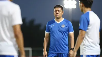 A 30-year-old male coach with a strong build, wearing a blue and white kit, standing on the sidelines, looking at the...