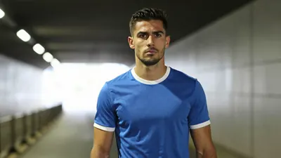 Mid-30s muscular midfielder, shoulders hunched, eyes fixed forward, wearing a plain blue shirt with white trim, soft...