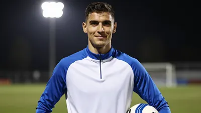 A focused, waist-up portrait of a 22-year-old forward with athletic build, short dark hair and intense eyes, wearing an...