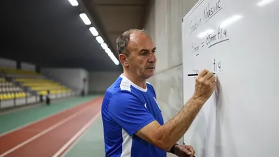 A 50-year-old male coach with a weathered face and receding hairline, wearing a plain blue and white polyester training...