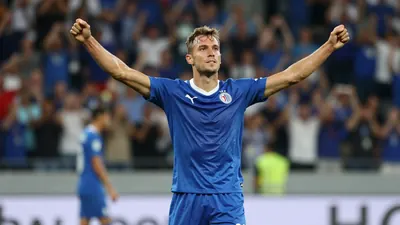 A muscular 25-30 year old Dinamo Zagreb midfielder in solid blue jersey and shorts, celebrating a goal with arms raised...