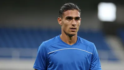 Mid-20s slender midfielder, dark hair swept back, intense gaze, wearing a plain blue training top with subtle white...