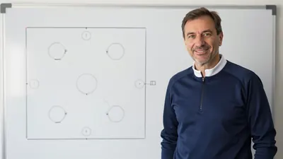 A photorealistic image of an anonymous GNK Dinamo coach in his mid-50s, with a kind face and a warm smile, standing in...