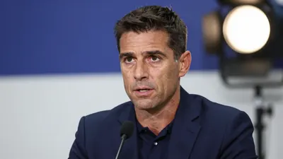 A head-and-shoulders portrait of a 40-45 year old male football coach in a dark blue suit, standing at a press conference...