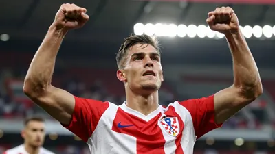 A muscular young Croatian footballer in a red and white striped jersey, celebrating a goal with arms raised, floodlit...