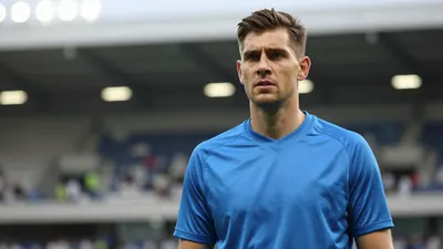 A 30-year-old goalkeeper with a muscular build, wearing a solid blue jersey, standing in a football stadium with...