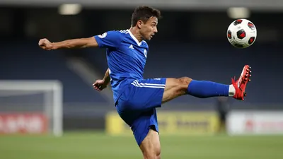 A muscular young striker in a blue jersey with a white stripe, mid-air during a bicycle kick, eyes focused on the ball