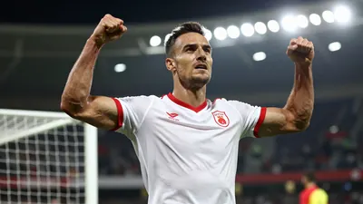 A muscular male football player in his late 20s, wearing a clean white jersey with red trim, celebrates a last-minute...