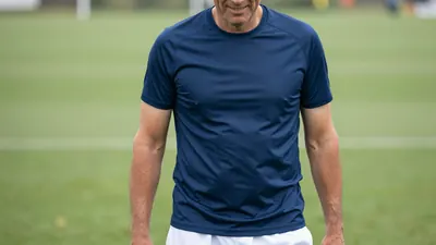 Middle-aged male club official, athletic build, waist-up, wearing a plain blue training shirt (solid navy fabric, subtle...
